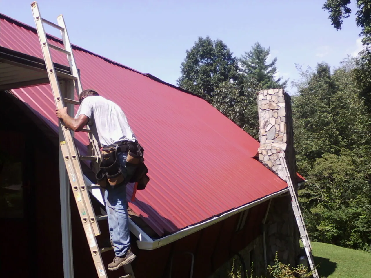 Roofer on a ladder beside a red metal roof and stone chimney — Soffit & Fascia Repair by Roofing Brothers in Eagan